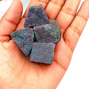 May include: Four rough-cut green and pink ruby gemstone crystals in a hand. The crystals are irregular in shape and have a natural, unpolished finish.