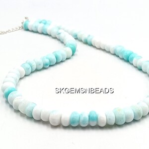 May include: A necklace composed of round, polished beads in shades of white and light blue. The beads are strung together, forming a necklace with a silver-colored clasp. The necklace is displayed on a white surface.