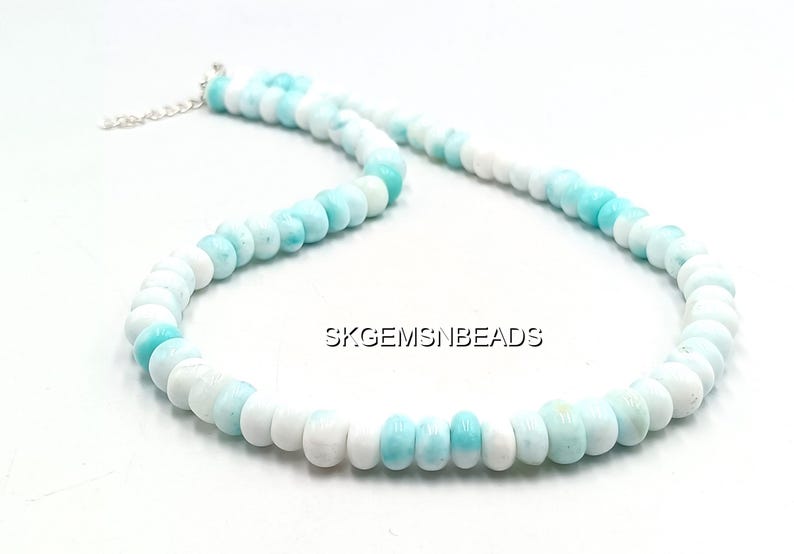 May include: A necklace composed of round, polished beads in shades of white and light blue. The beads are strung together, forming a necklace with a silver-colored clasp. The necklace is displayed on a white surface.