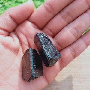 May include: Two black tourmaline crystals, one is a rectangular shape and the other is a long, thin shape. The crystals are in a person's hand.