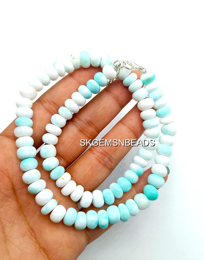 May include: A necklace composed of alternating white and light blue gemstone beads. The beads are oval-shaped and strung together with a silver clasp. The text "SKGEMSNBEADS" is visible in the image.