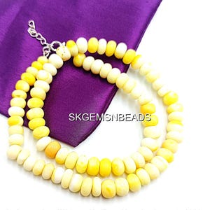 May include: A necklace made of cream and yellow beads, with a silver-colored clasp and extender chain. The beads are arranged in a circular pattern, resting on a purple cloth. The text "SKGEMSNBEDS" is visible.