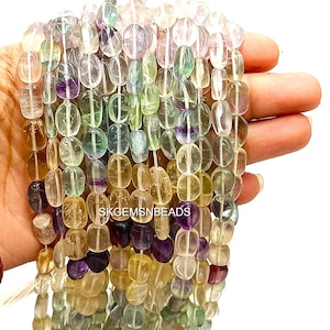 May include: Strands of colorful, oval-shaped gemstone beads in shades of purple, green, yellow, and clear. The beads are strung together, and the text "SKGEMSNBEADS" is visible. The beads are held in a hand against a white background.