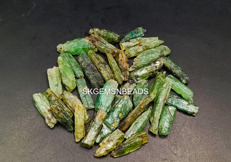 May include: A collection of green and brown rough gemstone points. The points are various sizes and shapes. The points are scattered on a black surface.