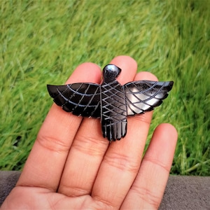 May include: A black carved bird figurine with outstretched wings. The bird has detailed wing and body patterns. The figurine is held in a hand against a green grassy background. The bird is a decorative item.