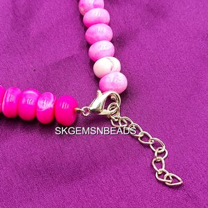 May include: A close-up of a pink beaded necklace with a gold-colored clasp and extender chain. The beads are a vibrant pink with white marbling. The text "SKGEMSNBEADS" is visible.