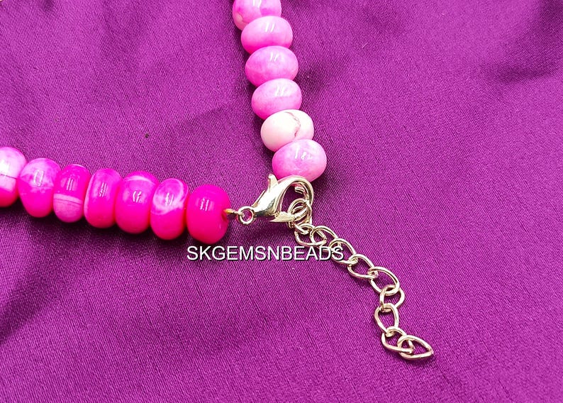 May include: A close-up of a pink beaded necklace with a gold-colored clasp and extender chain. The beads are a vibrant pink with white marbling. The text "SKGEMSNBEADS" is visible.