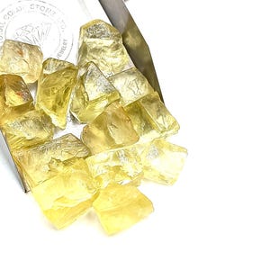 K&ouml;nnte beinhalten: Ein Haufen gelber, facettierter, roher Edelsteine. Die Steine befinden sich in einer Metallschale mit der Aufschrift "Natural Color Stone Co. Jewelry" auf der Seite.