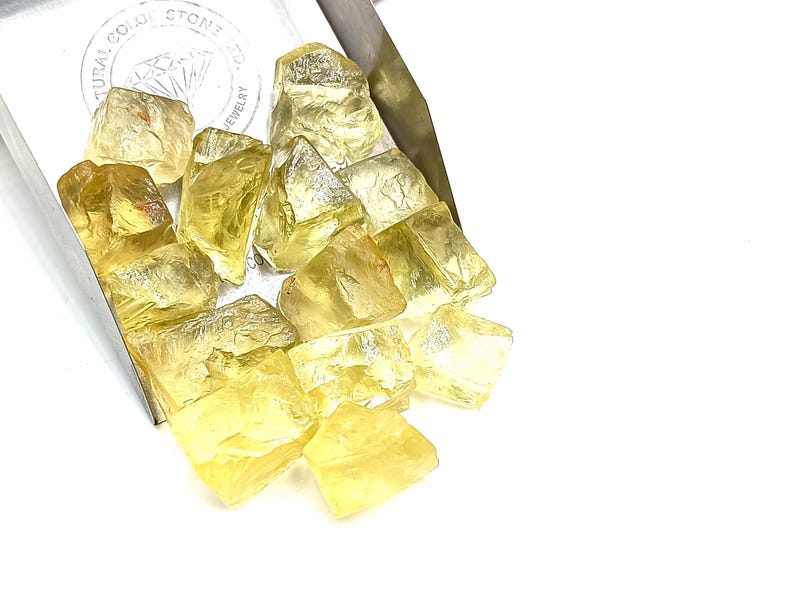 K&ouml;nnte beinhalten: Ein Haufen gelber, facettierter, roher Edelsteine. Die Steine befinden sich in einer Metallschale mit der Aufschrift "Natural Color Stone Co. Jewelry" auf der Seite.