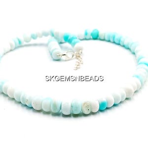 May include: A necklace composed of round, light blue and white beads. The beads are strung together, forming a circular shape. The necklace has a silver clasp. The text "SKGEMSNBEADS" is visible.