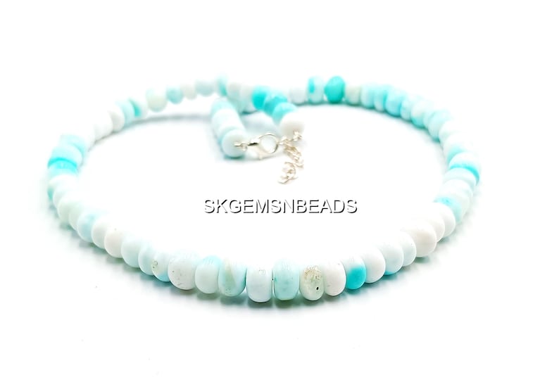 May include: A necklace composed of round, light blue and white beads. The beads are strung together, forming a circular shape. The necklace has a silver clasp. The text "SKGEMSNBEADS" is visible.