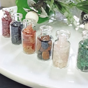 May include: Seven small glass bottles filled with various colored gemstones. The bottles are arranged in a row on a white surface.
