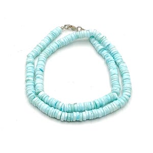 May include: A light blue beaded necklace with a silver clasp. The necklace is made of small, flat, round beads. The beads are a pale blue color, and the necklace is displayed on a white background.