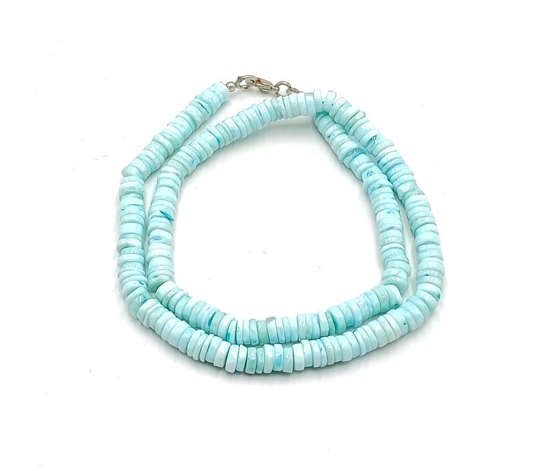 May include: A light blue beaded necklace with a silver clasp. The necklace is made of small, flat, round beads. The beads are a pale blue color, and the necklace is displayed on a white background.