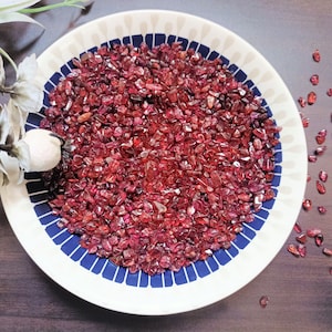 May include: A white ceramic plate with a blue and white pattern, filled with red ruby gemstone chips. The plate is on a dark brown wooden surface.