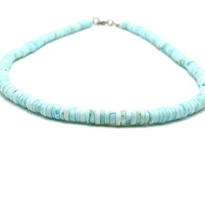 May include: A light blue beaded necklace with a silver clasp. The necklace is made of small, round, flat beads. The beads are a soft, pastel blue color, and the necklace is displayed against a white background.