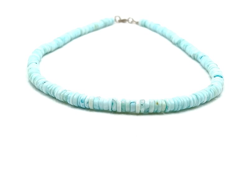 May include: A light blue beaded necklace with a silver clasp. The necklace is made of small, round, flat beads. The beads are a soft, pastel blue color, and the necklace is displayed against a white background.