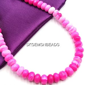 May include: A necklace of pink and white beads, with a gradient of colors from light to dark. The beads are round and smooth, resting on a purple fabric. The text "SKGEMSNBEADS" is visible.