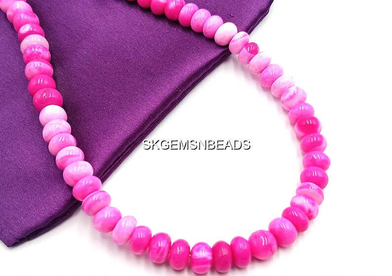 May include: A necklace of pink and white beads, with a gradient of colors from light to dark. The beads are round and smooth, resting on a purple fabric. The text "SKGEMSNBEADS" is visible.