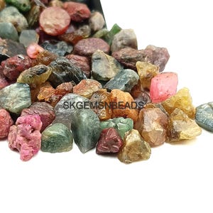 Pode incluir: Um monte de pedras preciosas de corte bruto coloridas, incluindo verde, azul, rosa e vermelho. As pedras preciosas estão espalhadas sobre uma superfície branca. SKGEMSNBEADS é visível na imagem.