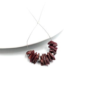 Può includere: Una fila di perline irregolari in pietra grezza color marrone scuro, intervallate da piccole perline distanziatrici sferiche in metallo color argento liscio, infilate su un filo metallico sottile, flessibile e color argento su sfondo bianco.