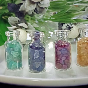 Puede incluir: Siete pequeñas botellas de vidrio llenas de piedras preciosas de varios colores. Las botellas están dispuestas en una fila sobre una superficie blanca.