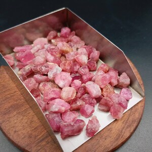 May include: A metal scoop filled with pink and red rough gemstones. The scoop is sitting on a wooden surface.