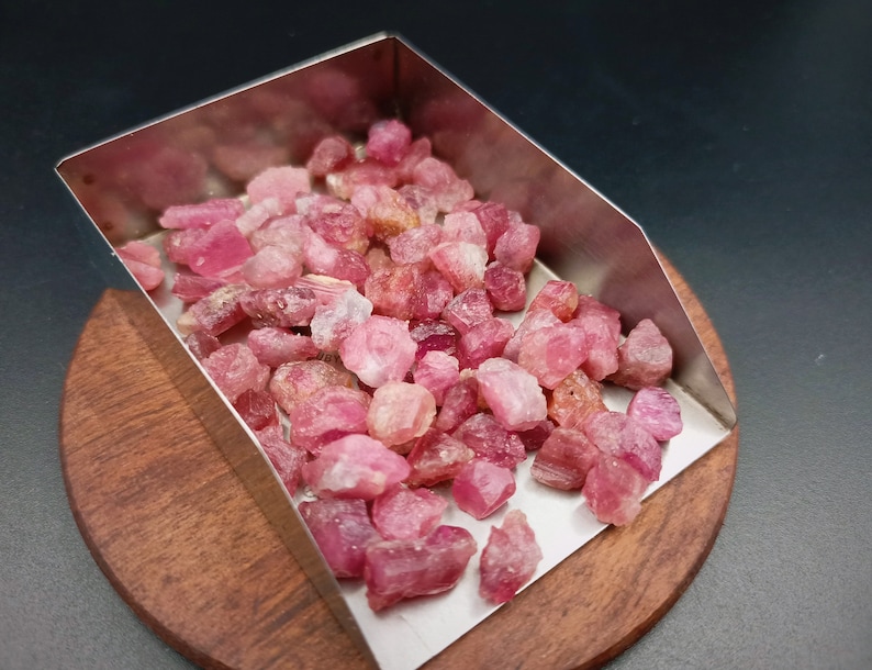 May include: A metal scoop filled with pink and red rough gemstones. The scoop is sitting on a wooden surface.