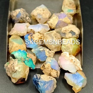 Puede incluir: Una colecci&oacute;n de piedras preciosas de &oacute;palo en bruto en varios tonos de azul, verde, rosa y blanco. Los &oacute;palos est&aacute;n colocados en una bandeja de metal. SKGEMSNBEADS est&aacute; escrito en la bandeja.