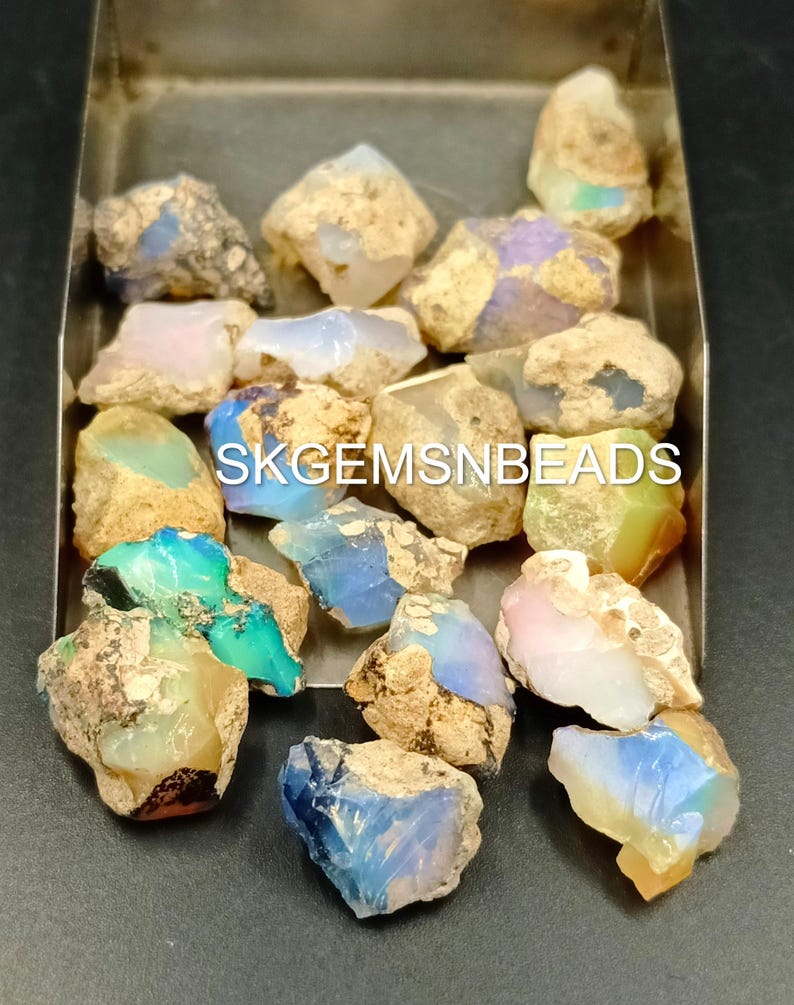 Puede incluir: Una colecci&oacute;n de piedras preciosas de &oacute;palo en bruto en varios tonos de azul, verde, rosa y blanco. Los &oacute;palos est&aacute;n colocados en una bandeja de metal. SKGEMSNBEADS est&aacute; escrito en la bandeja.