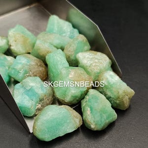 Op de afbeelding: Een verzameling lichtgroene en witte ruwe aquamarijn edelstenen in een metalen schep. De edelstenen zijn van verschillende groottes en vormen. SKGEMSNBEADS is op de schep gedrukt.