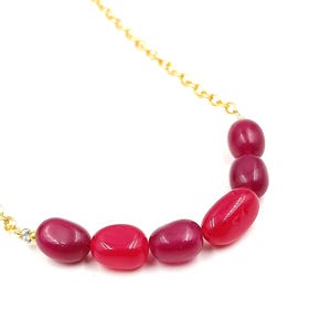 Op de afbeelding: Een gouden ketting met een rij van zes ovale rode en roze edelsteen kralen.