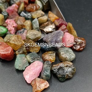 K&ouml;nnte beinhalten: Eine Sammlung von bunten, facettierten Edelsteinen, darunter rosa, gr&uuml;n, blau und braun, in verschiedenen Formen und Gr&ouml;&szlig;en. Die Edelsteine liegen lose und verstreut auf einer silbernen Oberfl&auml;che. Der Text "SKGEMSNBEADS" ist im Bild sichtbar.