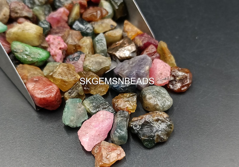 K&ouml;nnte beinhalten: Eine Sammlung von bunten, facettierten Edelsteinen, darunter rosa, gr&uuml;n, blau und braun, in verschiedenen Formen und Gr&ouml;&szlig;en. Die Edelsteine liegen lose und verstreut auf einer silbernen Oberfl&auml;che. Der Text "SKGEMSNBEADS" ist im Bild sichtbar.