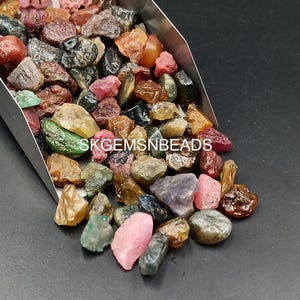 K&ouml;nnte beinhalten: Eine Schaufel mit bunten Edelsteinen, darunter rosa, rote, gr&uuml;ne, blaue und braune, in verschiedenen Formen und Gr&ouml;&szlig;en. Die Edelsteine liegen lose und verstreut in einer Metallschaufel. Der Text "SKGEMSNBeads" ist im Bild sichtbar.