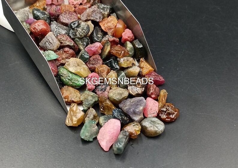 K&ouml;nnte beinhalten: Eine Schaufel mit bunten Edelsteinen, darunter rosa, rote, gr&uuml;ne, blaue und braune, in verschiedenen Formen und Gr&ouml;&szlig;en. Die Edelsteine liegen lose und verstreut in einer Metallschaufel. Der Text "SKGEMSNBeads" ist im Bild sichtbar.
