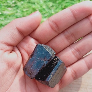 May include: A dark brown, almost black, crystal with a rough, natural texture. The crystal is shaped like a cube and has a slightly iridescent sheen.