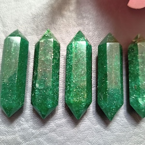 May include: Six green crystal points with a sparkly, iridescent finish. The points are arranged in a row on a light gray surface.