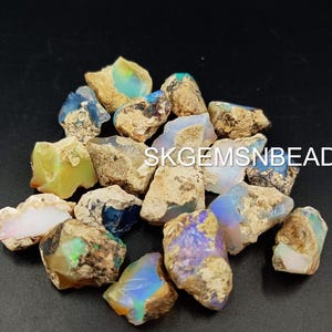 Puede incluir: Una colecci&oacute;n de piedras preciosas de &oacute;palo en bruto en varios tonos de azul, verde y blanco. Los &oacute;palos est&aacute;n engastados en una matriz de roca marr&oacute;n y gris. El texto "SKGEMSNBEADS" es visible en la imagen.