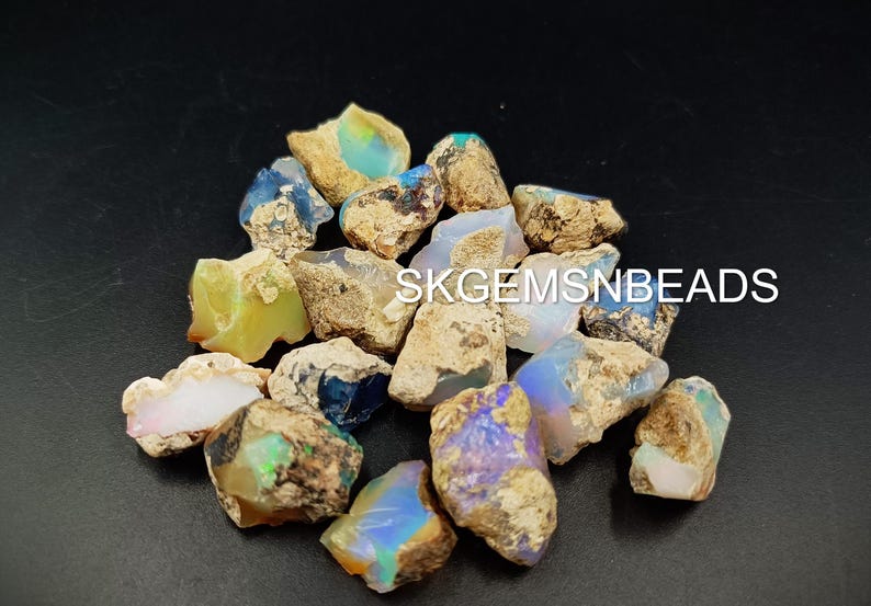 Puede incluir: Una colecci&oacute;n de piedras preciosas de &oacute;palo en bruto en varios tonos de azul, verde y blanco. Los &oacute;palos est&aacute;n engastados en una matriz de roca marr&oacute;n y gris. El texto "SKGEMSNBEADS" es visible en la imagen.
