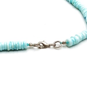 May include: A close-up of a light blue beaded necklace with a silver clasp. The necklace features small, round, flat beads. The clasp is a simple, secure design. The necklace is on a white background.