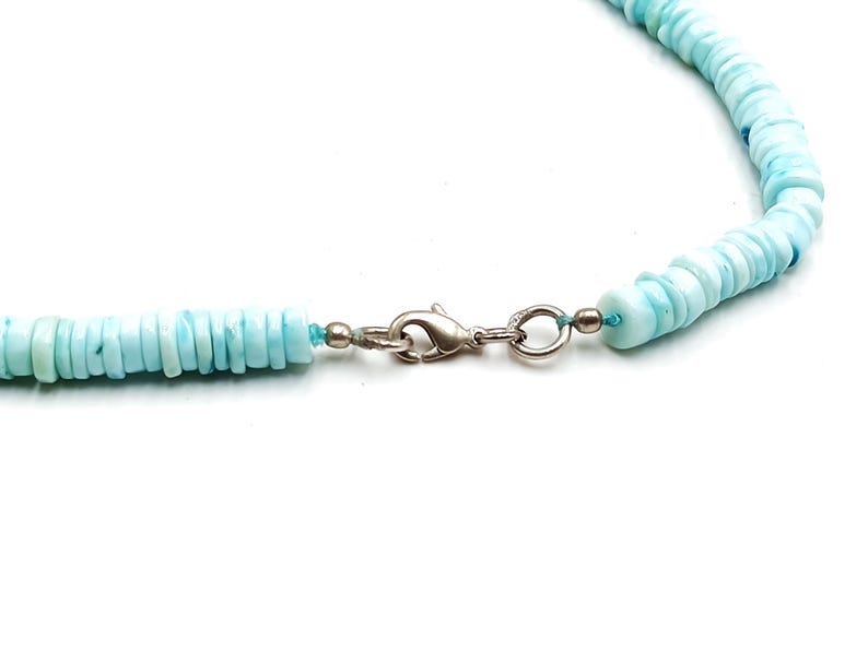May include: A close-up of a light blue beaded necklace with a silver clasp. The necklace features small, round, flat beads. The clasp is a simple, secure design. The necklace is on a white background.