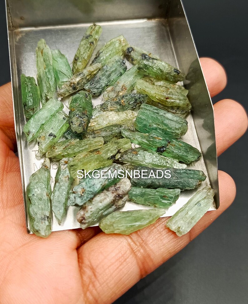 May include: A collection of green and black gemstone rough, also known as green apatite, in a metal tray. The rough is in various sizes and shapes. The rough is ready to be cut and polished into gemstones.