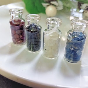 Könnte beinhalten: Vier kleine Glasflaschen gefüllt mit verschiedenen farbigen Edelsteinen. Die Flaschen sind in einer Reihe auf einer weißen Oberfläche angeordnet. Die Edelsteine sind rot, blau, weiß und dunkelblau.