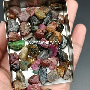 K&ouml;nnte beinhalten: Eine Sammlung von rohen, ungeschliffenen Edelsteinen in verschiedenen Farben, darunter Rot, Gr&uuml;n, Schwarz und Gelb. Die Edelsteine befinden sich in einer kleinen Metallschale. Der Text "SKGEMSNBEADS" ist auf der Schale sichtbar.