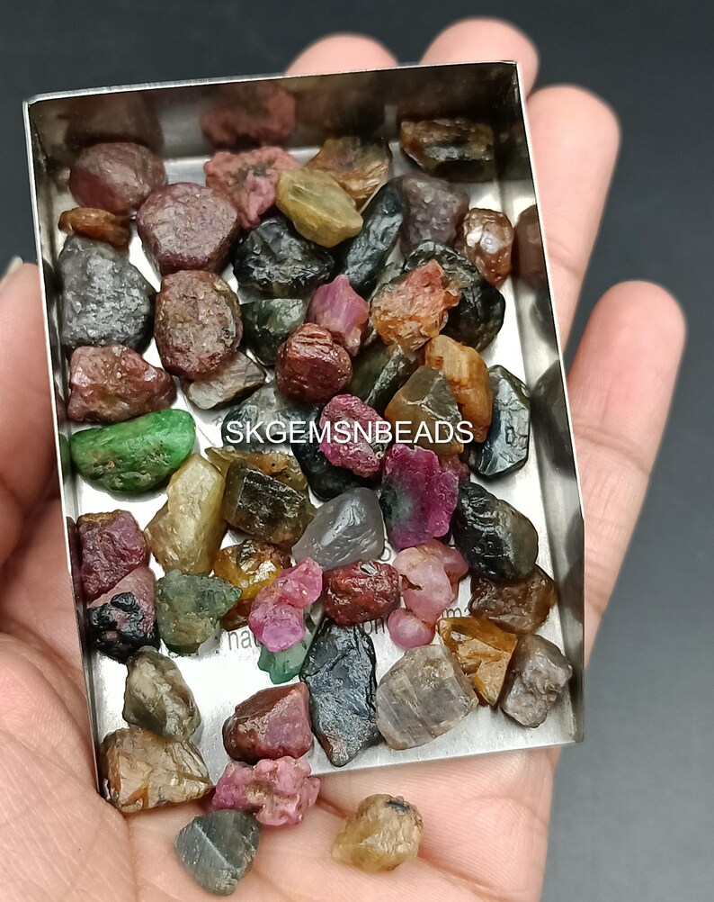 K&ouml;nnte beinhalten: Eine Sammlung von rohen, ungeschliffenen Edelsteinen in verschiedenen Farben, darunter Rot, Gr&uuml;n, Schwarz und Gelb. Die Edelsteine befinden sich in einer kleinen Metallschale. Der Text "SKGEMSNBEADS" ist auf der Schale sichtbar.