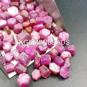 May include: A close-up of a pile of pink ruby gemstone rough. The rough is in various sizes and shapes. The rough is in a metal scoop. The text "SKGEMSNBEADS" is visible in the image.