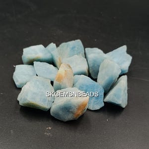 May include: A collection of rough-cut aquamarine gemstones. The stones are various sizes and shapes, with a pale blue color and some with tan and brown patches. The gemstones are scattered on a dark surface.