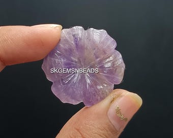 May include: A carved amethyst gemstone in the shape of a flower. The gemstone is a light purple color with carved petal details. The text "SKGEMSNBEADS" is visible on the gemstone.