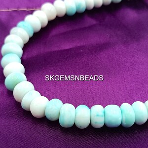 May include: A necklace of round, light blue and white beads. The beads are strung together, creating a curved shape against a purple background. The text "SKGEMSNEADS" is visible.
